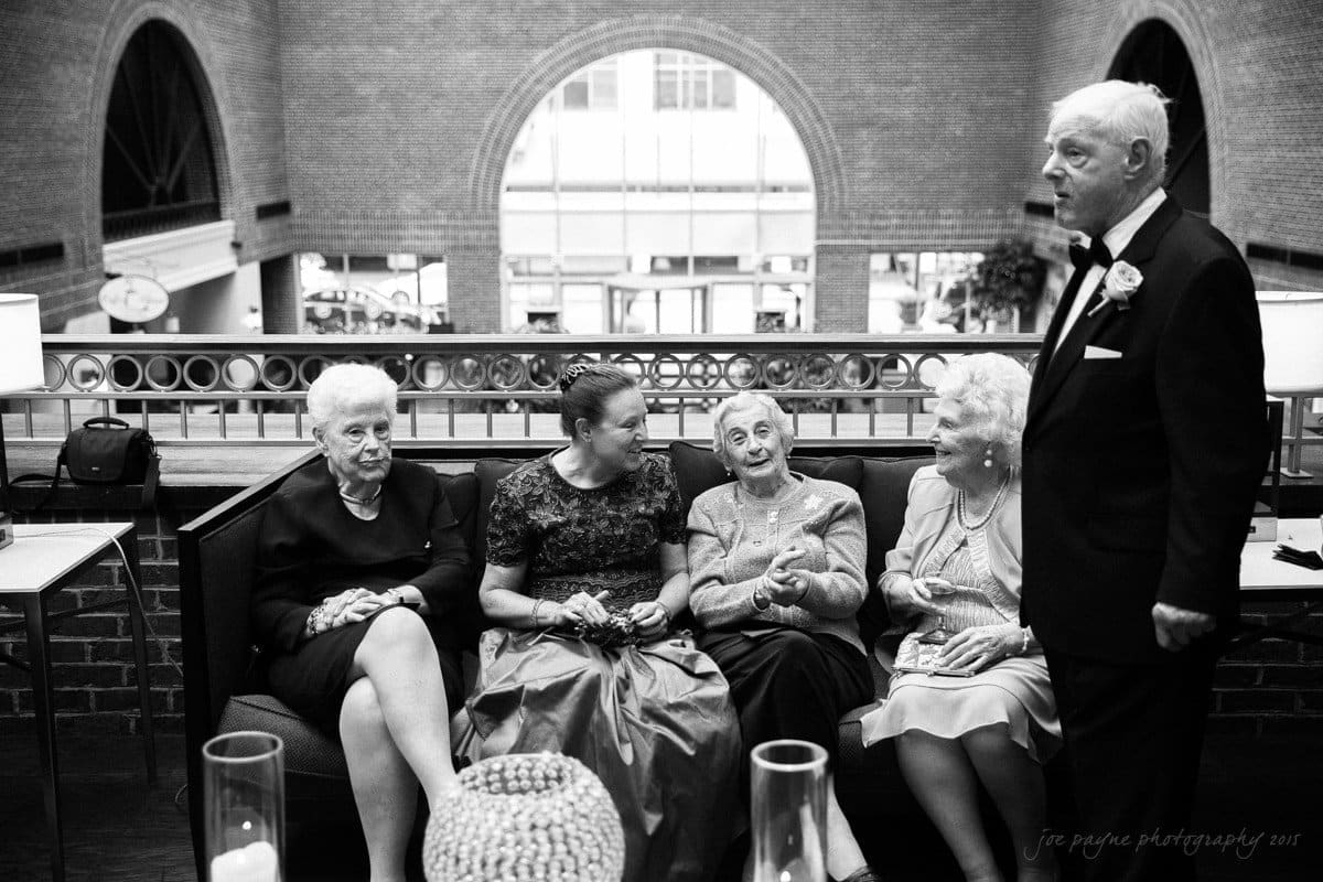 A black and white photo of four elderly women seated on a sofa, engaged in conversation. A man in a suit stands to the right. The setting is a grand room with arched windows and brick walls, suggesting a formal gathering or event.