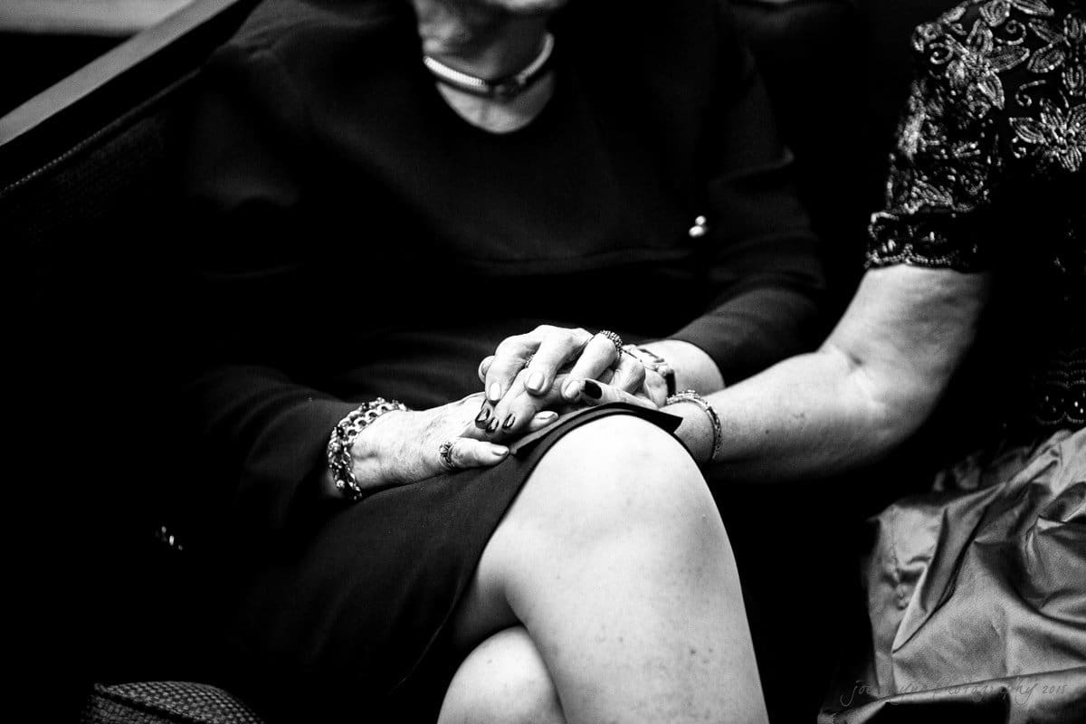 Two people sitting closely, with one person resting their hand on the others. Both are wearing dark clothing, and the photo is in black and white, emphasizing the textures and patterns of their outfits.