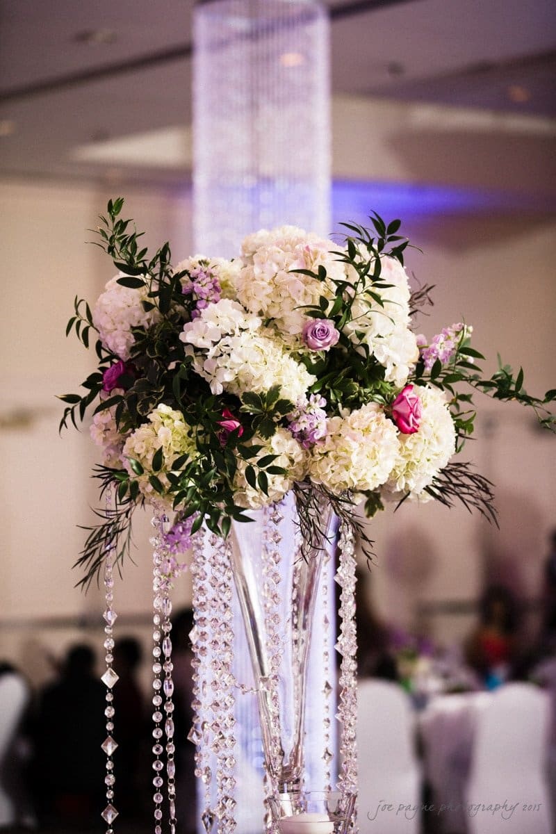 A tall glass vase filled with white hydrangeas, pink and purple roses, and greenery is adorned with hanging crystal strands. The floral arrangement stands prominently in a softly lit room, providing an elegant and luxurious centerpiece.