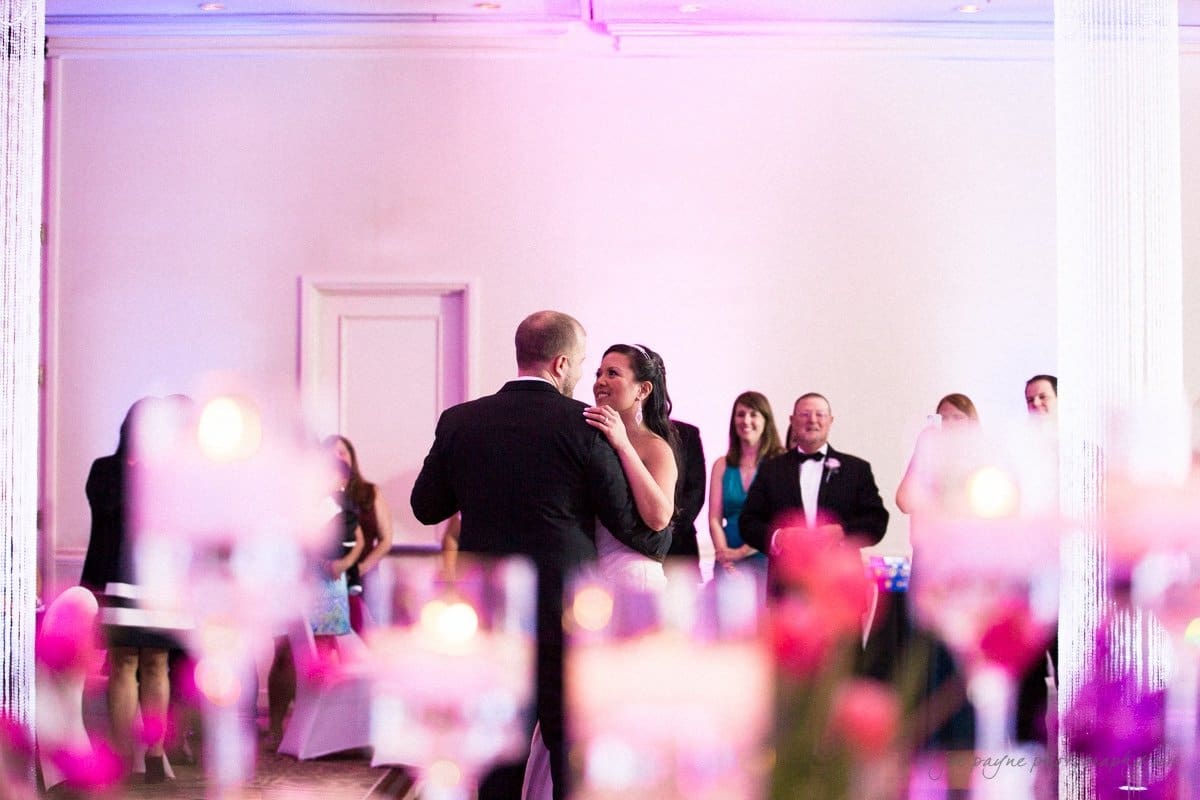 A couple dances in a softly lit room adorned with pink and purple lighting. The foreground is blurred with candles and flowers, while a group of people in formal attire watches from the background.