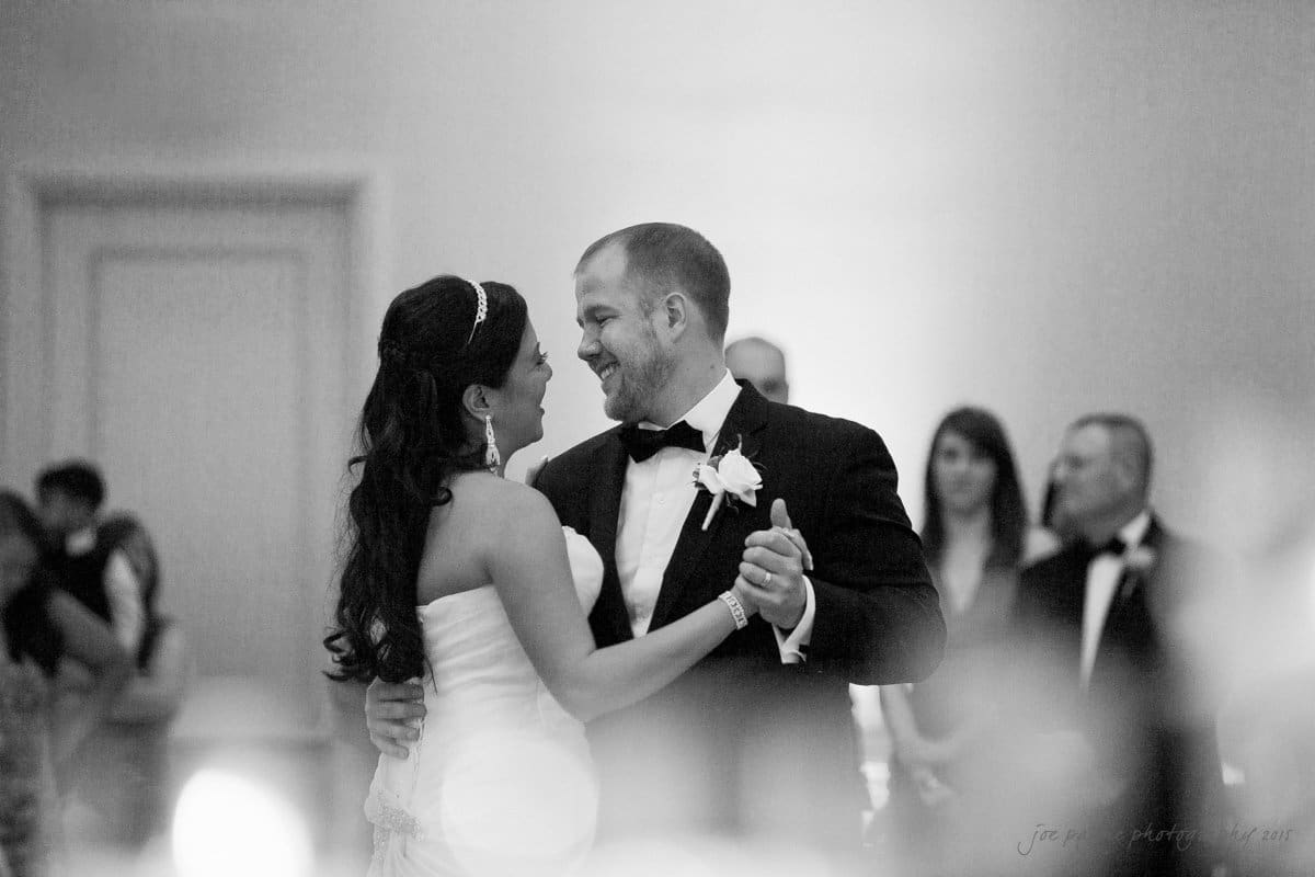 Bride and groom smiling at each other while dancing in a formal setting. The bride is wearing a strapless gown and the groom is in a tuxedo. A few guests are visible in the blurred background.