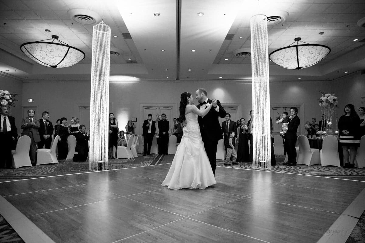 A bride and groom share a first dance in a large, elegant ballroom. Guests stand around the dance floor, watching. The room features modern lighting and decorative columns, creating an intimate and celebratory atmosphere.