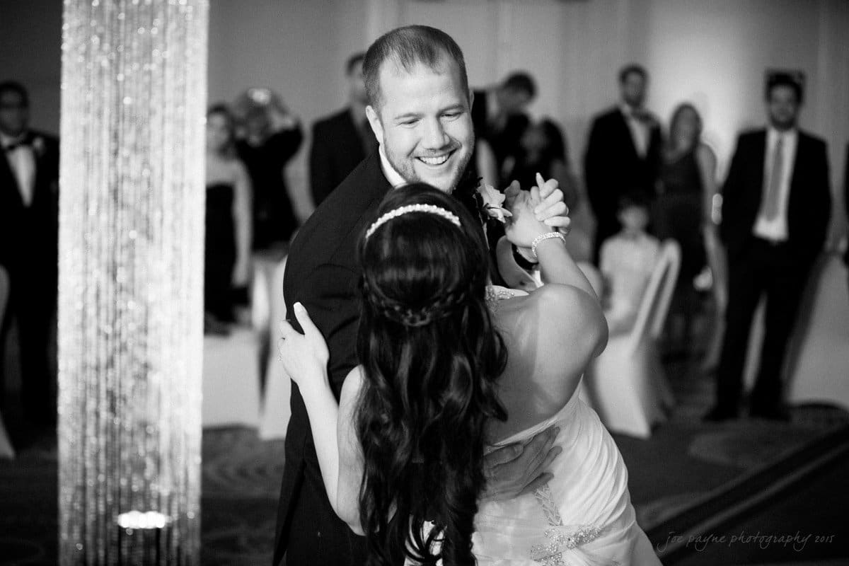 A bride and groom share their first dance at their wedding. The groom is smiling as the bride, wearing a strapless gown and hairpiece, embraces him. Guests watch in the softly lit ballroom in the background.
