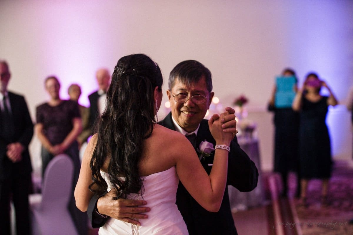 A bride in a white dress dances with an older man wearing a suit in a warmly lit room. Guests in the background watch and take photos, with some clapping. The scene feels joyful and celebratory.