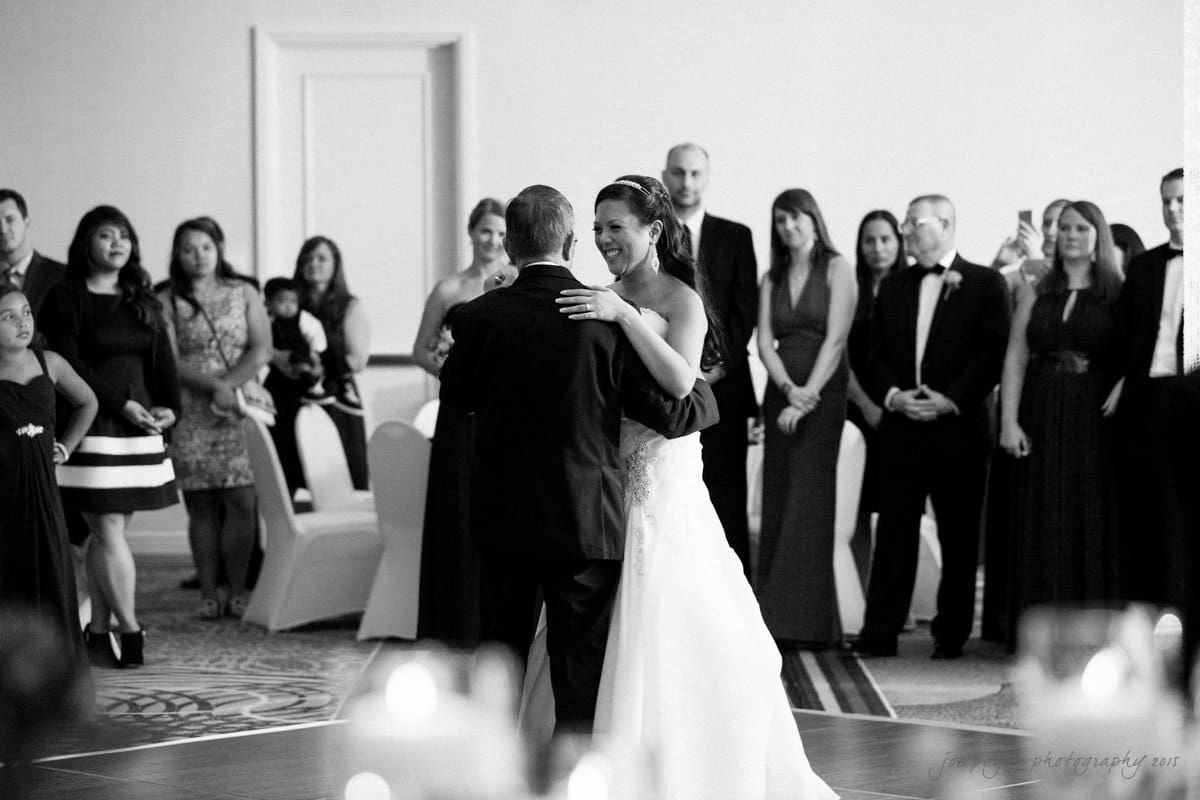 A bride and an older man share a dance on a ballroom floor surrounded by elegantly dressed guests. The setting is formal, with guests watching and smiling. Candles and chairs are visible, adding to the intimate atmosphere.
