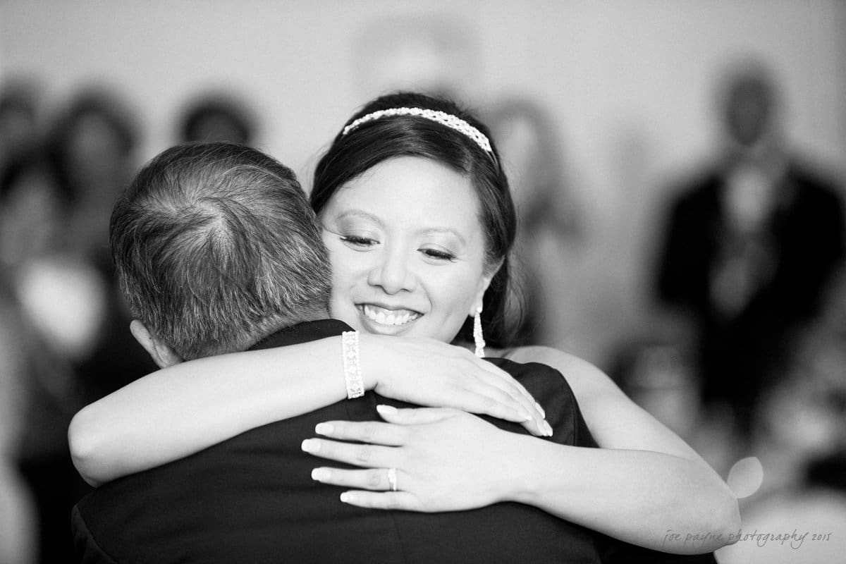 A happy bride with a tiara hugs an older man in a suit, smiling warmly. The background is softly blurred, suggesting a wedding reception atmosphere.