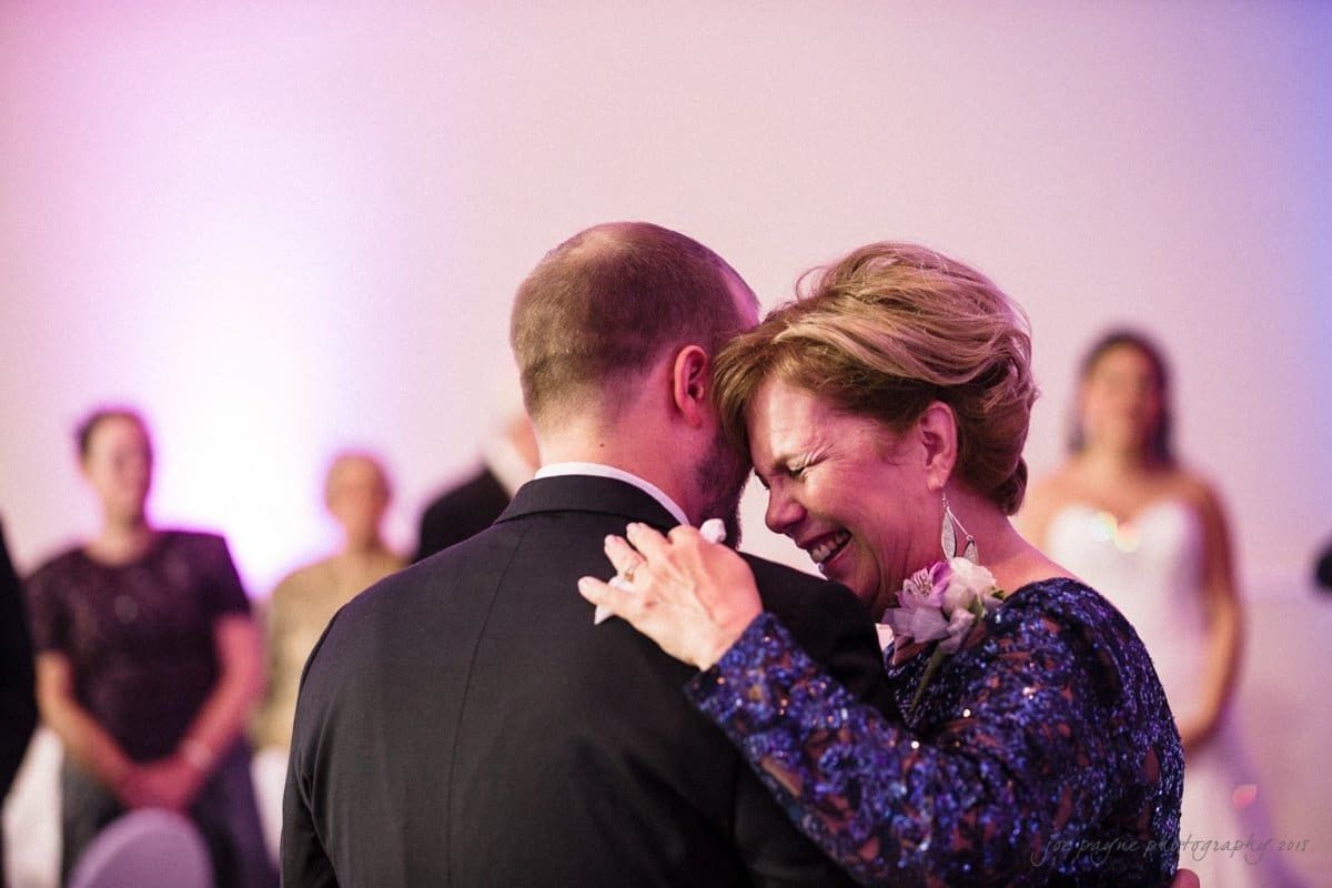 A woman and a man share a joyful moment while dancing at an event. The woman is dressed in a sparkling blue outfit with a flower corsage, and both appear to be smiling. People in the background watch them.