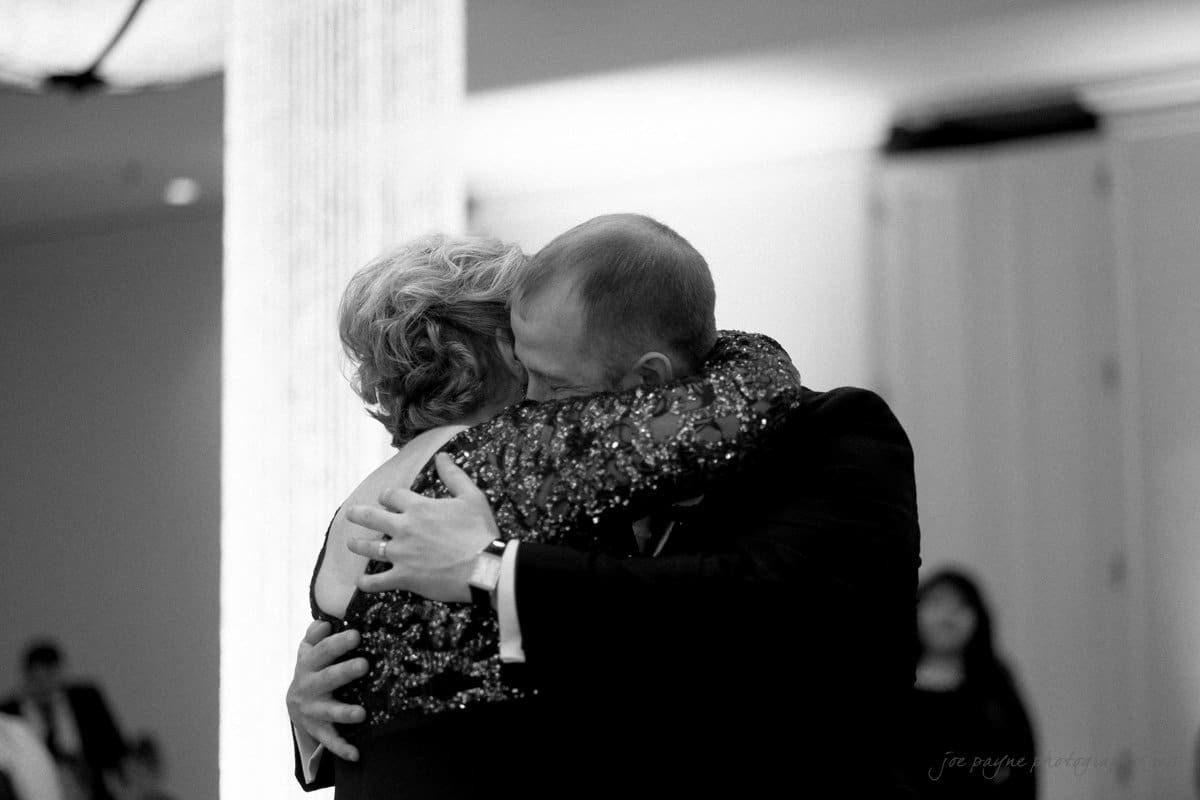 A black and white photo of an older woman and a man hugging tightly, surrounded by soft indoor lighting. The woman is wearing a sequined dress, and both appear emotional, perhaps at a significant event. A blurred figure is visible in the background.