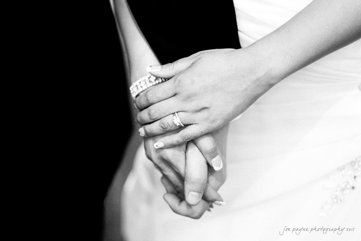 Close-up of a couple holding hands, with focus on a womans hand wearing a wedding ring. The background is dark, highlighting the white dress and the hands, suggesting a wedding or formal occasion.