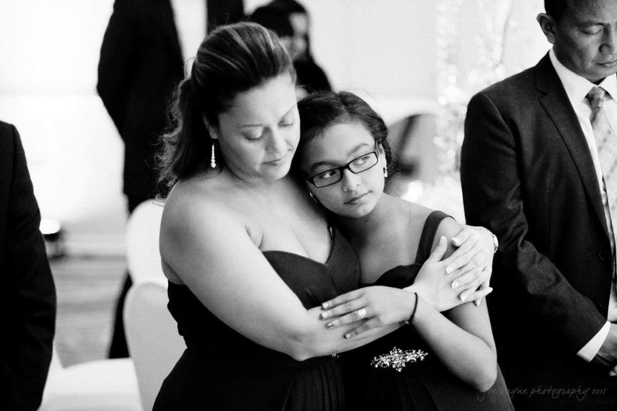 A black-and-white photo of a woman and a young girl hugging closely, both wearing formal dresses. The woman has her eyes closed, and the girl is looking slightly to the side. A man in a suit is visible in the background.