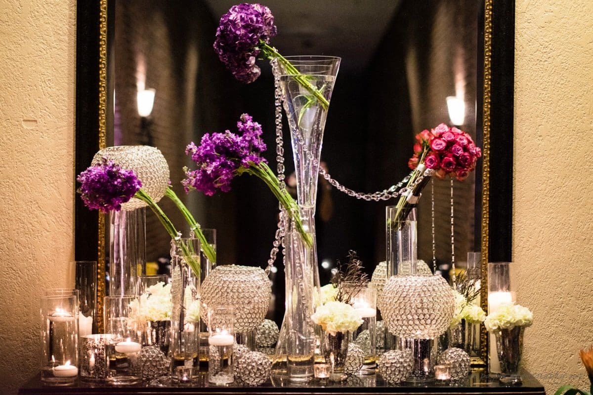 An elegant floral arrangement featuring tall glass vases with purple and pink flowers, surrounded by smaller white blooms and decorative crystal ornaments. Lit candles add a warm ambiance, reflecting on a mirror in the background.