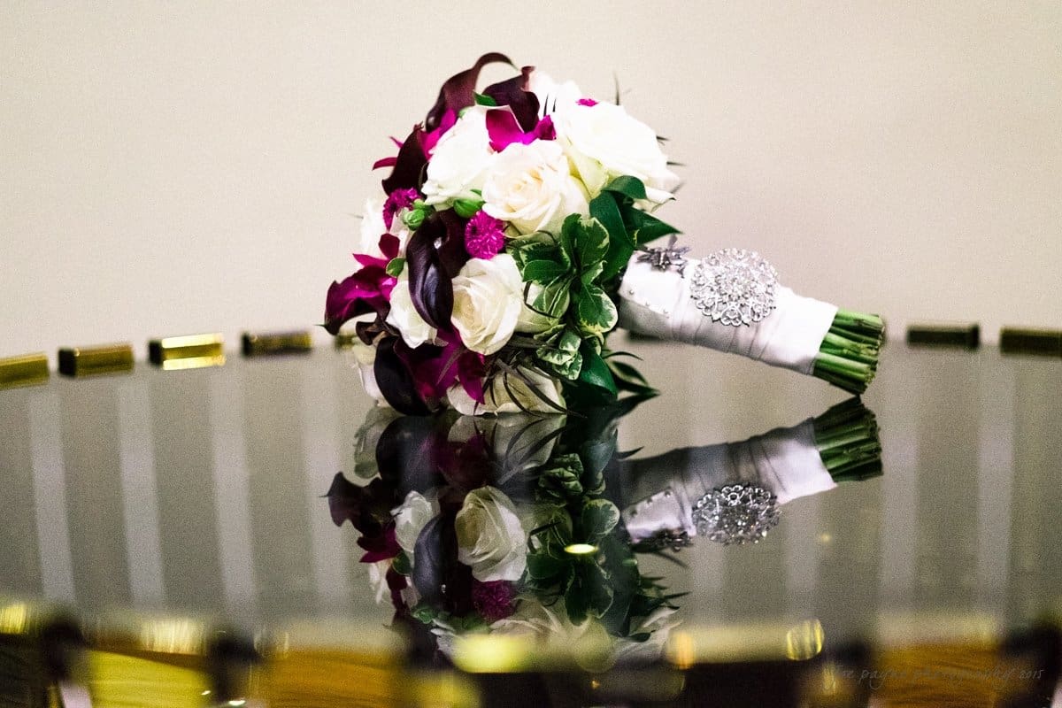 A bouquet of white, pink, and purple flowers with green leaves is wrapped in white fabric adorned with a jeweled brooch. It rests on a reflective surface with a gold-edged border, creating a mirror image of the bouquet.