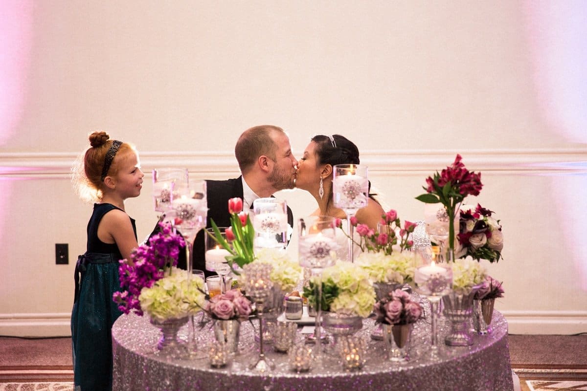 A couple shares a kiss at a wedding reception. They sit behind a round table adorned with colorful flowers and candles. A young girl in a blue dress and headband smiles beside them. The background is softly lit in pink and white.