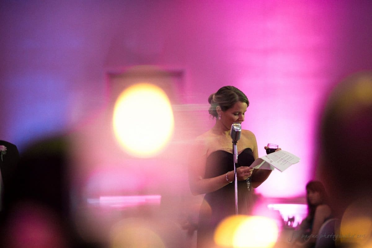 A woman in a black dress stands at a microphone, holding a piece of paper and a glass. The background is lit in pink and purple hues, creating a festive atmosphere. Defocused lights appear in the foreground.