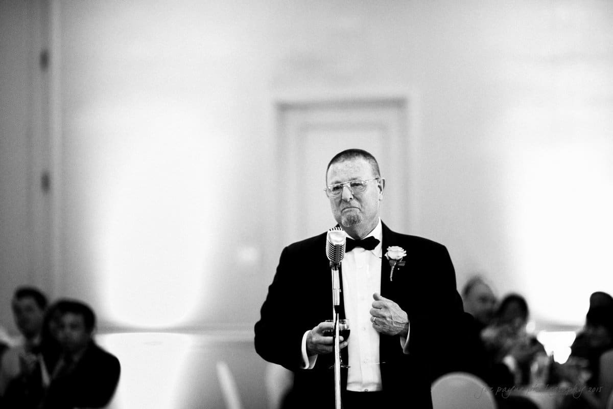 A man in a suit and bow tie stands in front of a vintage microphone, giving a speech. The background is blurred, with seated people listening attentively. The image is in black and white.