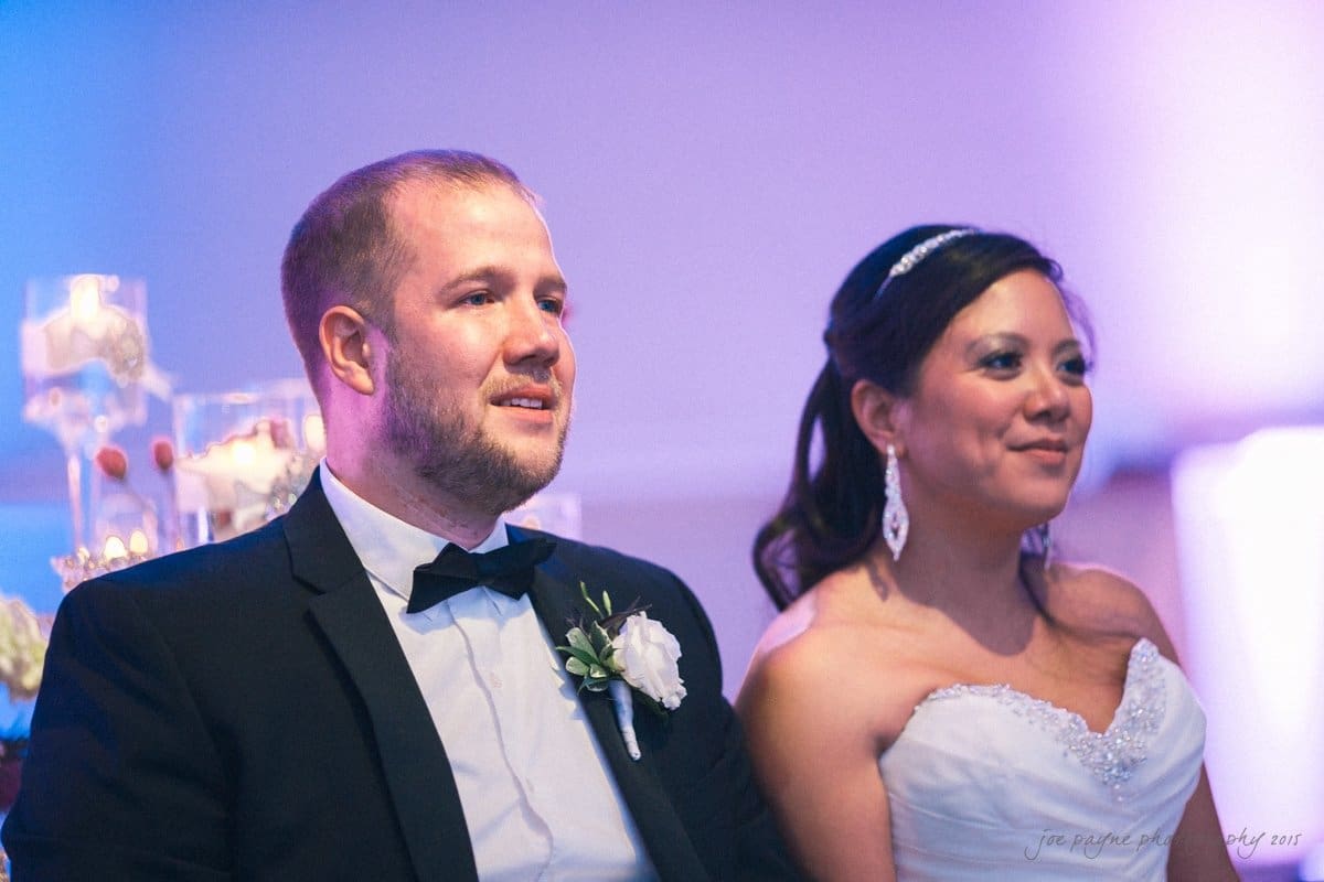A couple sits together, smiling. The man is wearing a black tuxedo with a bow tie and a white boutonniere. The woman is in a white strapless gown with a jeweled necklace and earrings. They are in a softly lit setting.