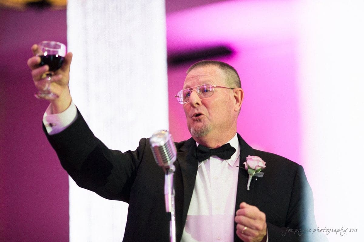 A man in a tuxedo holds a glass of red wine aloft, making a toast. He stands in front of a vintage microphone, with a pink-lit background.