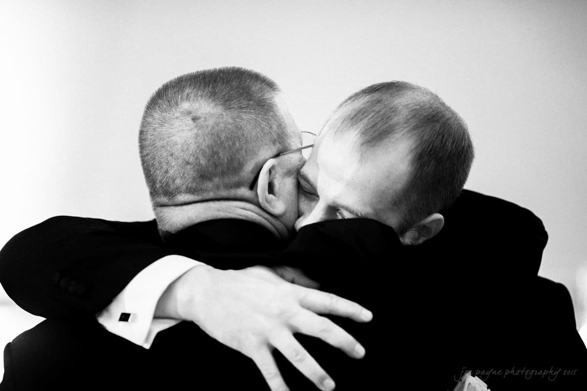 Two men in suits embrace warmly, their faces close together, conveying a sense of deep emotion and connection. The background is softly blurred, and the lighting is gentle, highlighting the personal moment between them.