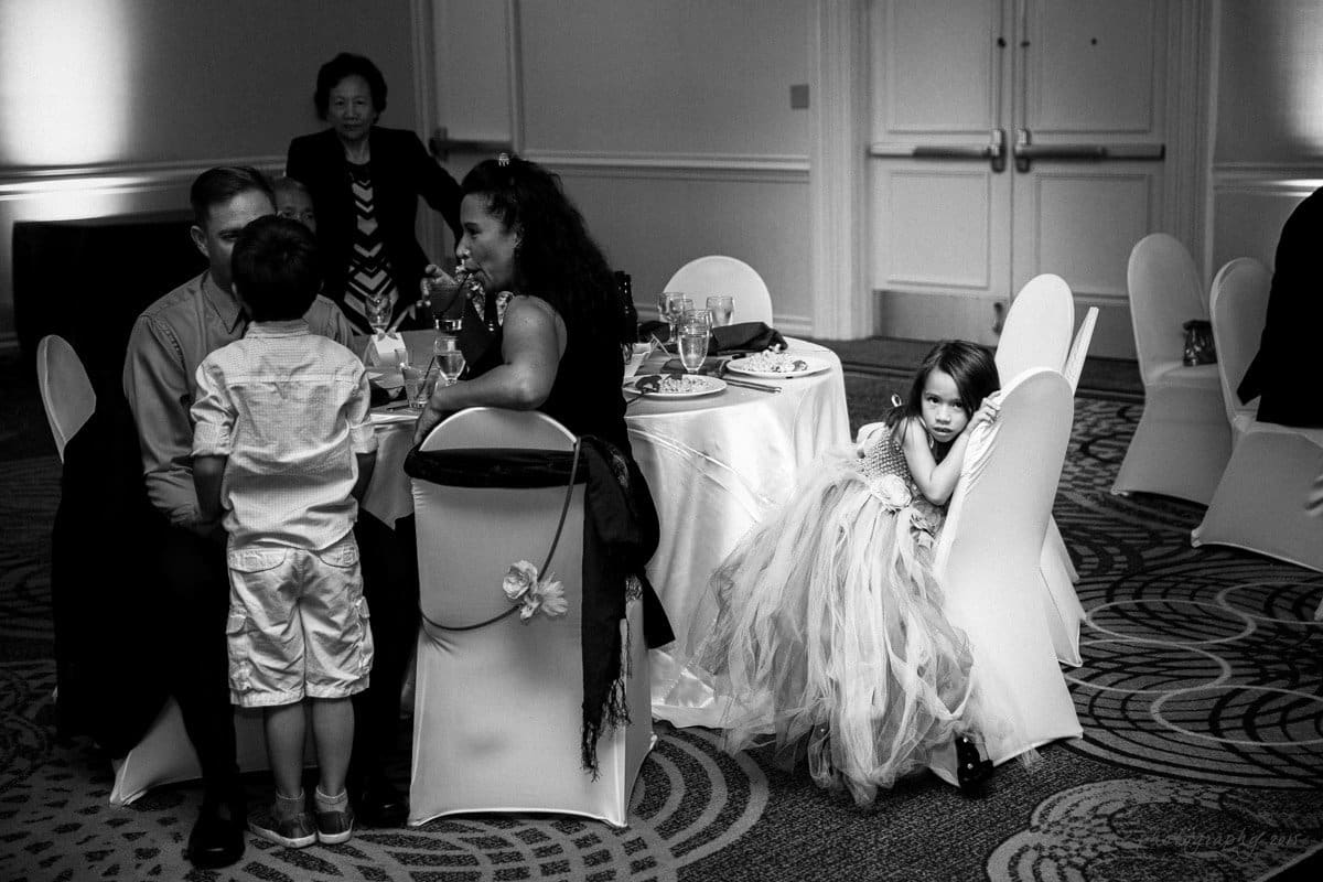 A black-and-white photo of a formal event. A young girl in a tutu dress sits on a chair, resting her head on her arms. Nearby, a couple with a child gather around a table with plates and drinks. A woman stands in the background by double doors.