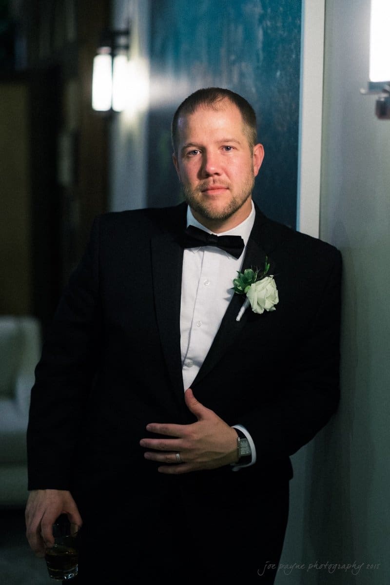 A man in a tuxedo with a bow tie stands against a wall, holding a drink in his right hand. He is wearing a boutonniere with white flowers on his lapel. The setting is dimly lit, creating a formal and elegant atmosphere.