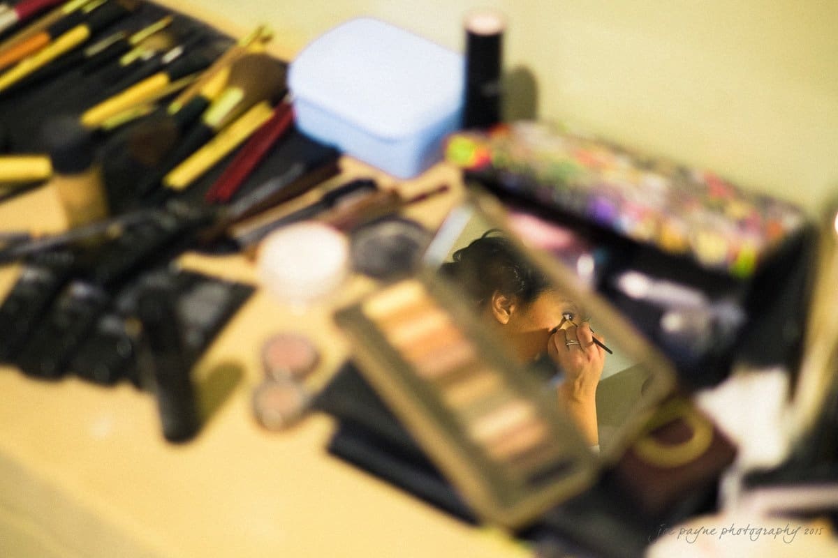 A close-up of a makeup table with various brushes, palettes, and cosmetics scattered. A woman is reflected in a small mirror applying mascara. The scene is brightly lit, emphasizing the vibrant colors of the makeup products.