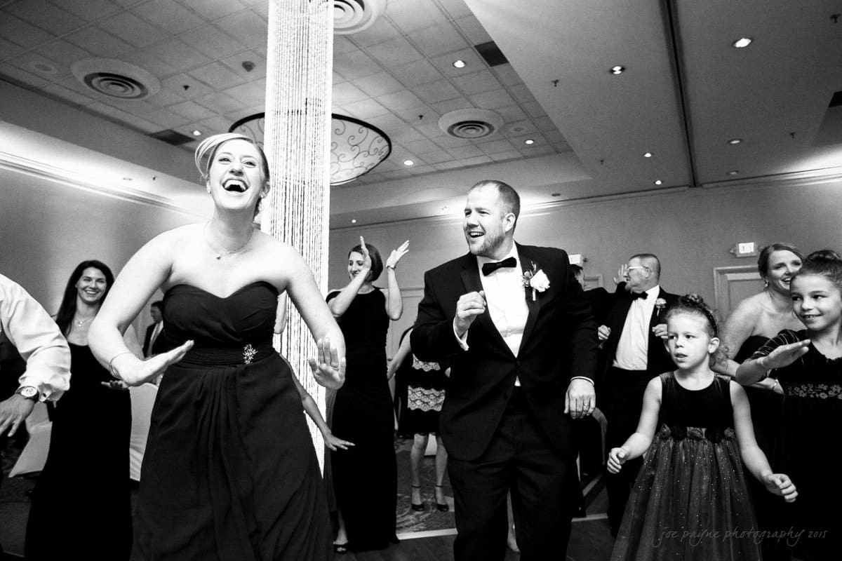 A joyful bride in a strapless gown and groom in a tuxedo dance enthusiastically at a wedding reception. Guests surround them, laughing and clapping. Two young girls in dresses join the fun on the dance floor. The atmosphere is festive and lively.