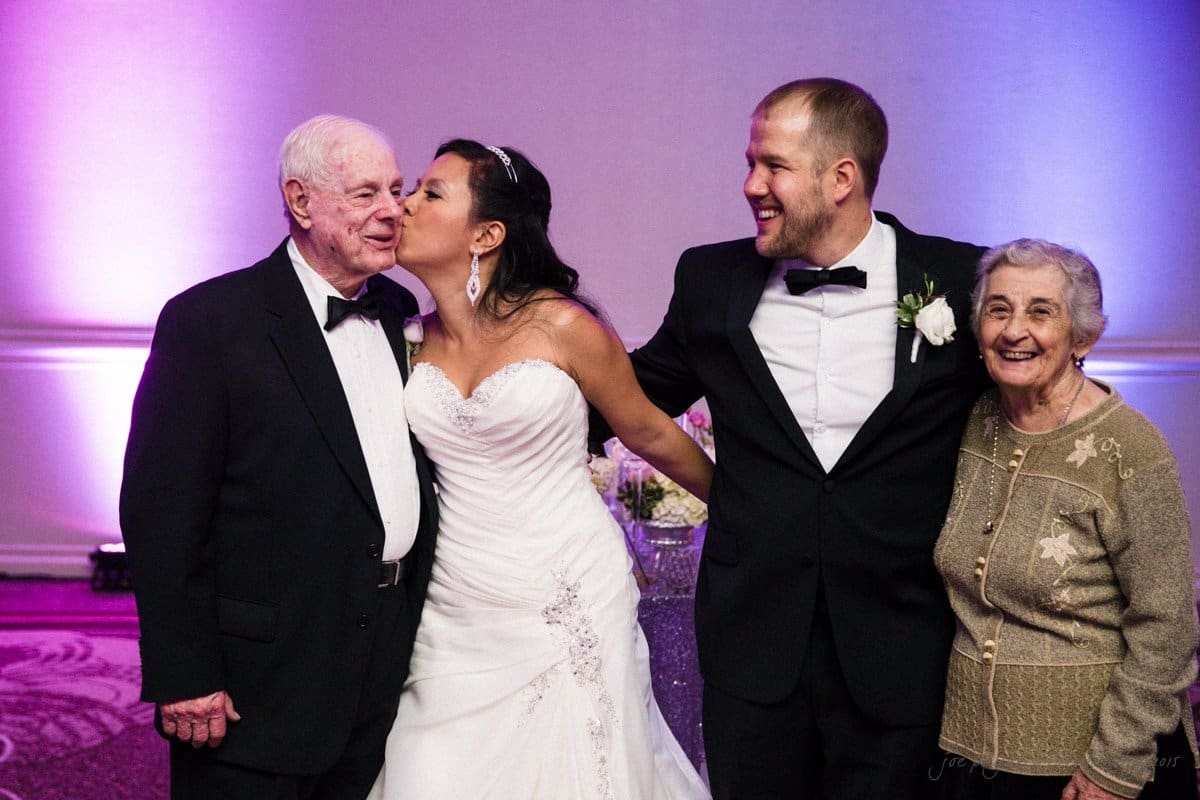 A bride in a white dress kisses an elderly man in a black suit on the cheek. Next to them, a groom in a black tuxedo stands arm-in-arm with an elderly woman in a beige sweater. They are gathered together, smiling in a warmly lit room.