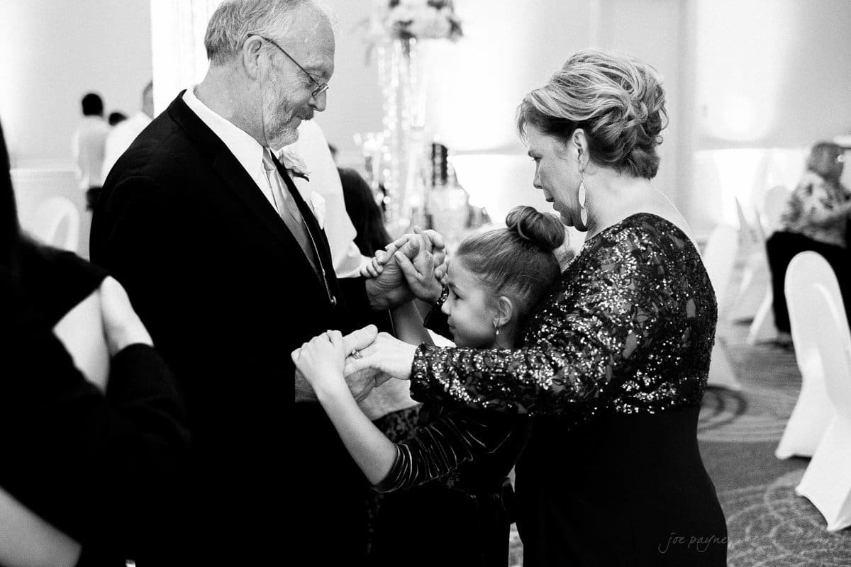 A man in a suit and a woman in a sequined dress dance with a young girl at a formal event. They hold hands, smiling and interacting joyfully in a well-lit room with white chairs and tables in the background.