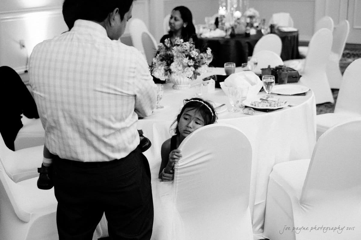 A young girl wearing a headband peeks from behind a chair at a banquet. A man in a striped shirt stands nearby. Tables are set with flowers and glasses, and a woman sits in the background. The scene is in black and white.