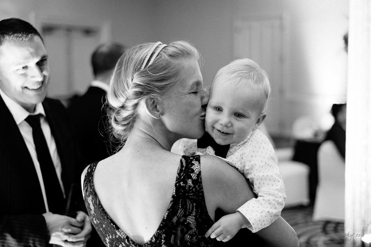 A woman in a formal dress is holding a smiling baby, gently kissing their cheek. A man in a suit stands nearby, smiling warmly. The scene takes place indoors at a social event. Black and white photo.