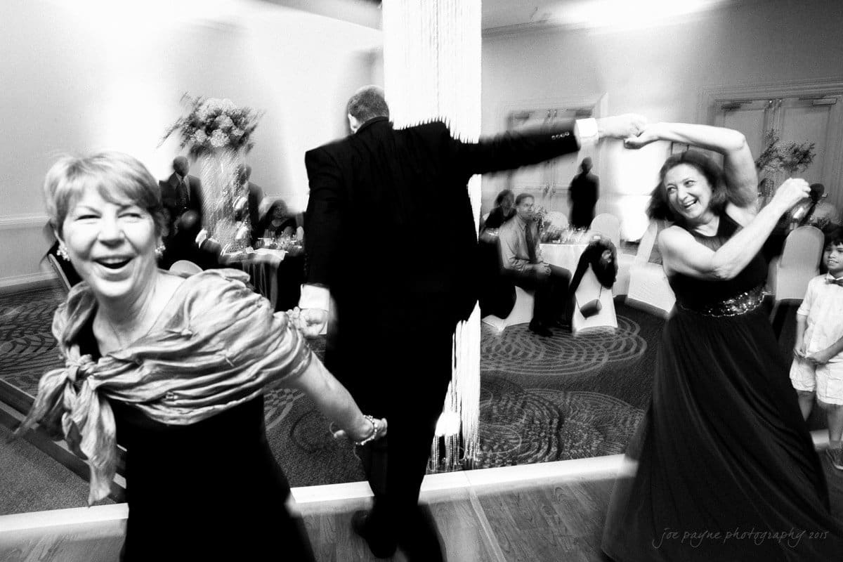 A lively black-and-white photo of people dancing joyfully at a wedding reception. A woman in a shawl smiles while holding hands with a dancing man. Guests in the background are sitting and enjoying the celebration.
