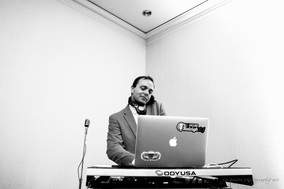 A DJ stands at a booth with a laptop and equipment. He is wearing headphones around his neck and is dressed in a suit. A microphone is positioned to the left of the setup. The scene is in black and white.