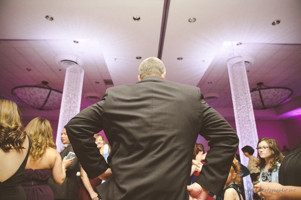 A man in a suit stands with his back to the camera, hands on hips, at a formal event. People around him are engaged in conversation, and the room is decorated with columns and purple lighting.