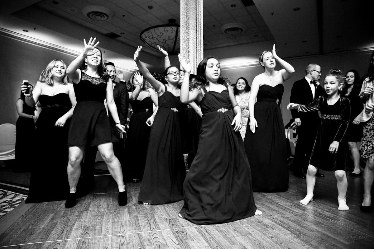 A group of people in formal attire are dancing energetically on a wooden floor in a ballroom setting. Women in dresses and a few children enjoy the lively atmosphere, with one person holding a drink. The mood is joyful and festive.