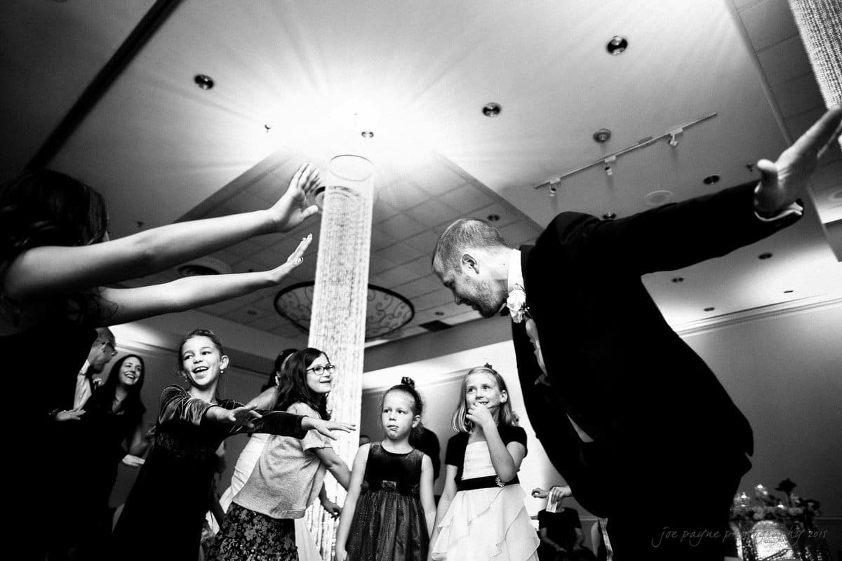 A man in a suit dances energetically in a ballroom, surrounded by smiling children. The children are dressed in formal attire, and the scene captures a joyful moment with a bright light overhead.