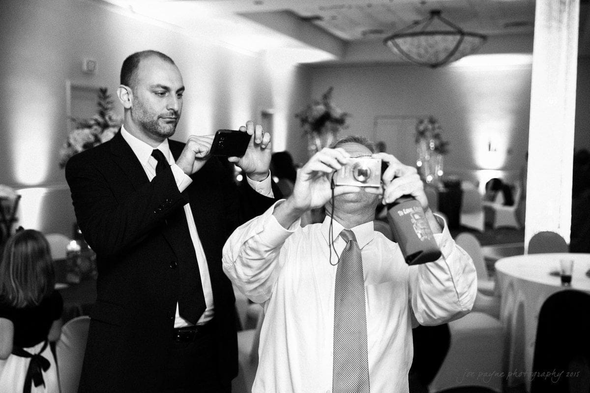 Two men in suits taking photos at an event. One man is holding a smartphone while the other uses a digital camera. The scene is set in a decorated room with people in the background. Black and white photo.