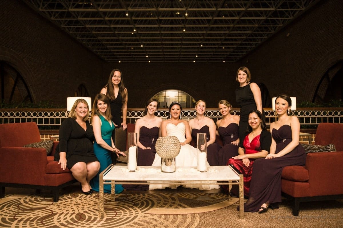 A bride sits on a couch surrounded by her bridesmaids in a formal setting. The women are dressed in elegant gowns and are smiling. There are decorative vases on the table in front of them. The background features an arched window and ceiling pattern.