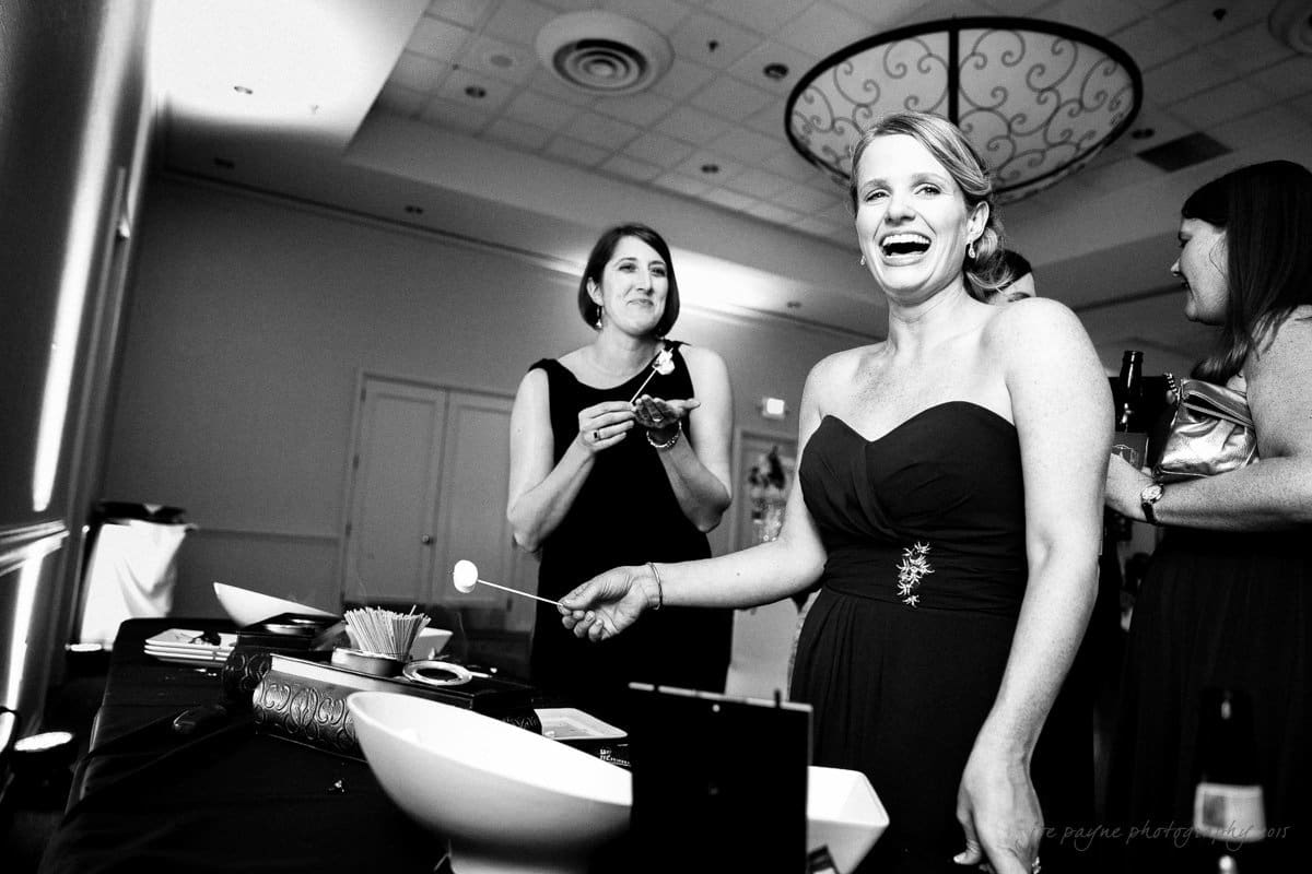 A black and white photo of three women at a social event. The woman in the foreground is smiling and holding a marshmallow on a stick. Two other women stand nearby, one holding a plate. There are bowls and a marshmallow roasting setup on the table.