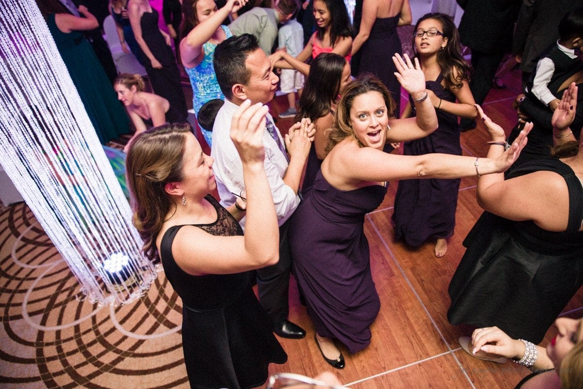 A lively group of people dancing at a party. They are dressed in formal attire, and the dance floor is vibrant with energy. A woman in a purple dress is in the center, raising her hands. The background is filled with colorful lights.