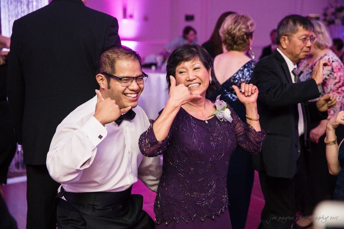 Two people smiling and making a playful hand gesture as if holding a phone. They are dressed in formal attire at a lively event with other guests mingling in the background.