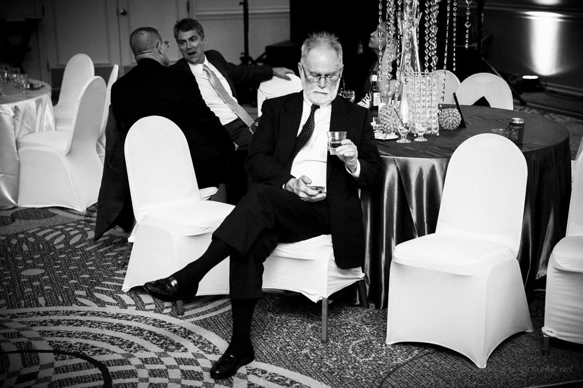 Black and white photo of a formal event. An older man with white hair and a beard sits holding a drink, engrossed in his phone. In the background, two men in suits are engaged in conversation at a round table with chairs covered in white fabric.