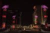A nighttime cityscape featuring four tall, illuminated structures with pink and red lights lining a wide pathway. Modern buildings flank the street on both sides, and soft lights add a serene atmosphere.