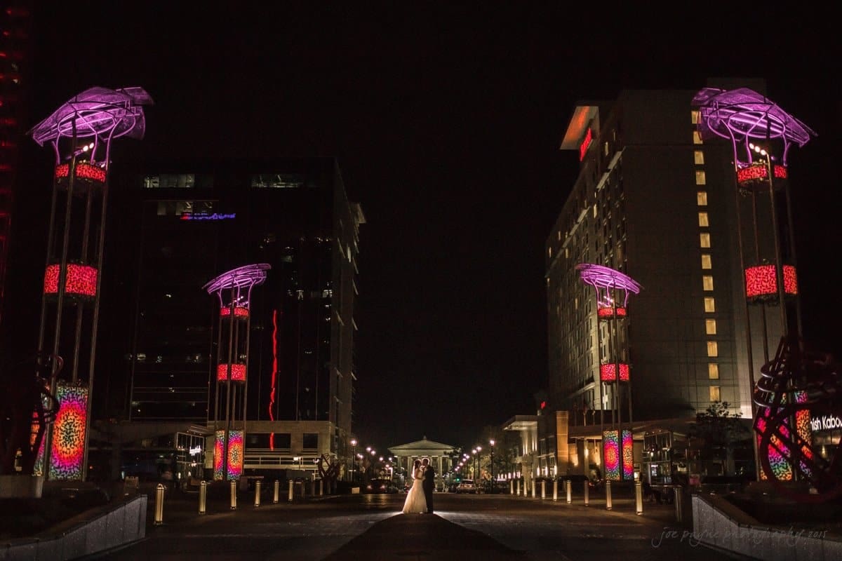 A nighttime cityscape featuring four tall, illuminated structures with pink and red lights lining a wide pathway. Modern buildings flank the street on both sides, and soft lights add a serene atmosphere.