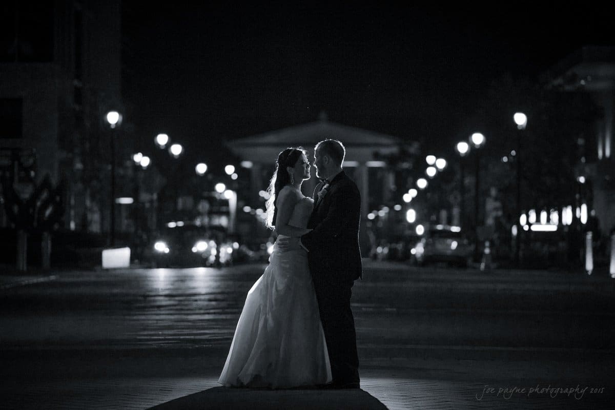 A couple in formal attire stands closely together on a street at night, illuminated by streetlights. The background is blurred, highlighting their silhouetted figures and romantic ambiance.