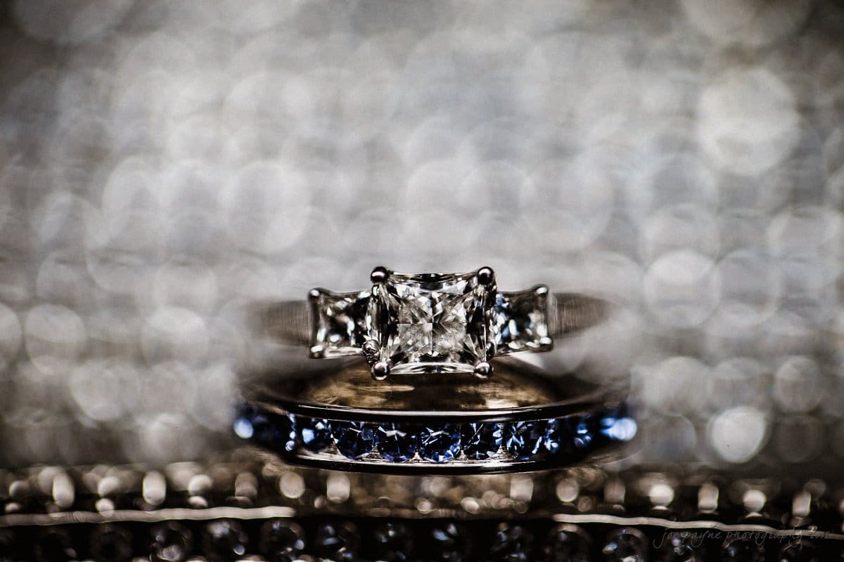 Close-up of a set of rings with a central diamond and two smaller side diamonds set in a silver band. Below is another silver band adorned with blue stones, both against a blurred, bokeh background.