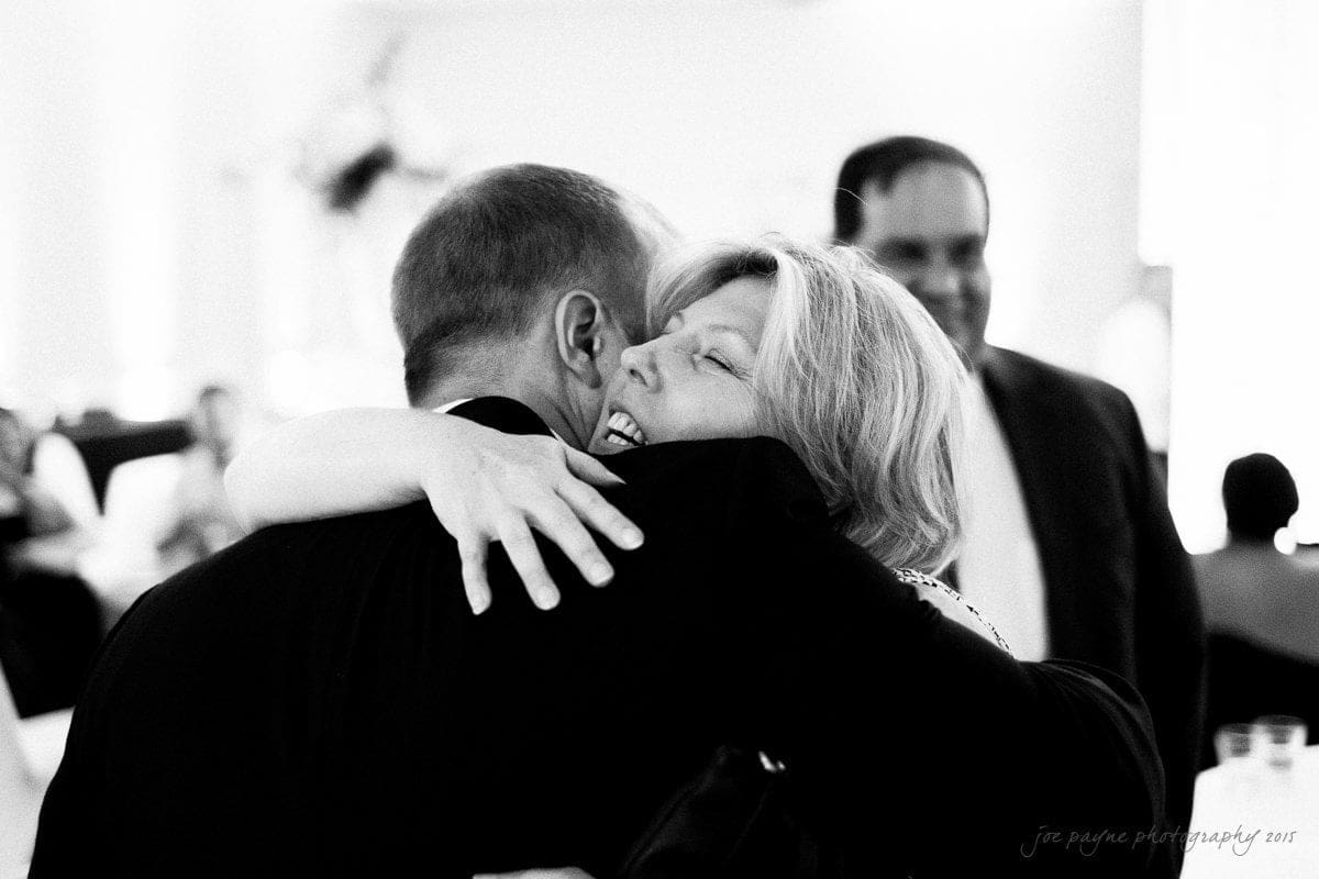 A woman joyfully hugs a man at a gathering, with another man smiling in the background. The setting appears to be a social event or celebration.