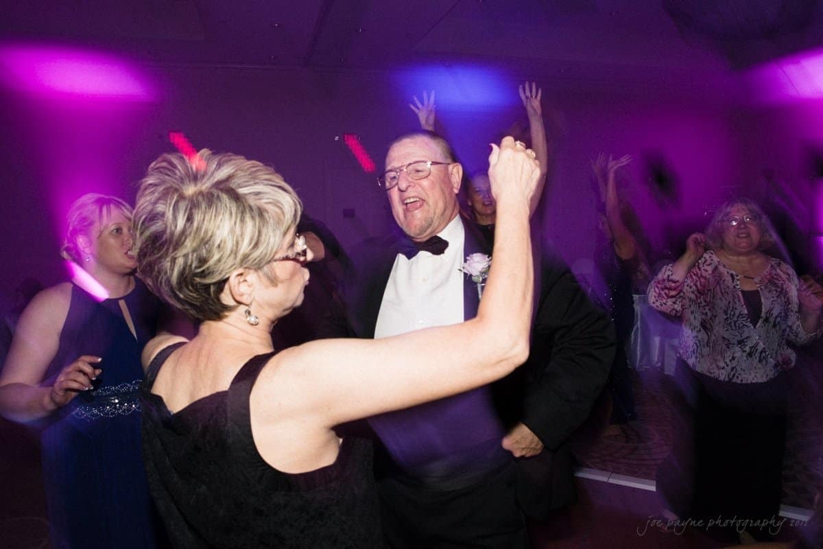 A couple joyfully dancing in formal attire at a party. The man is wearing a tuxedo, and the woman is in a black dress. They are surrounded by other guests under vibrant purple lighting.