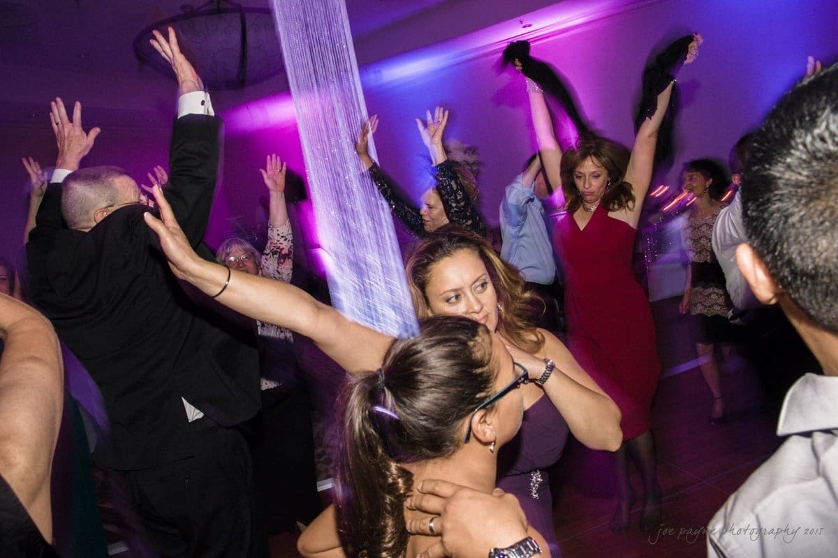 A lively dance floor with people in formal attire joyfully dancing. A woman in a red dress and another in a black suit raise their arms, surrounded by vibrant purple lighting and energetic motion.