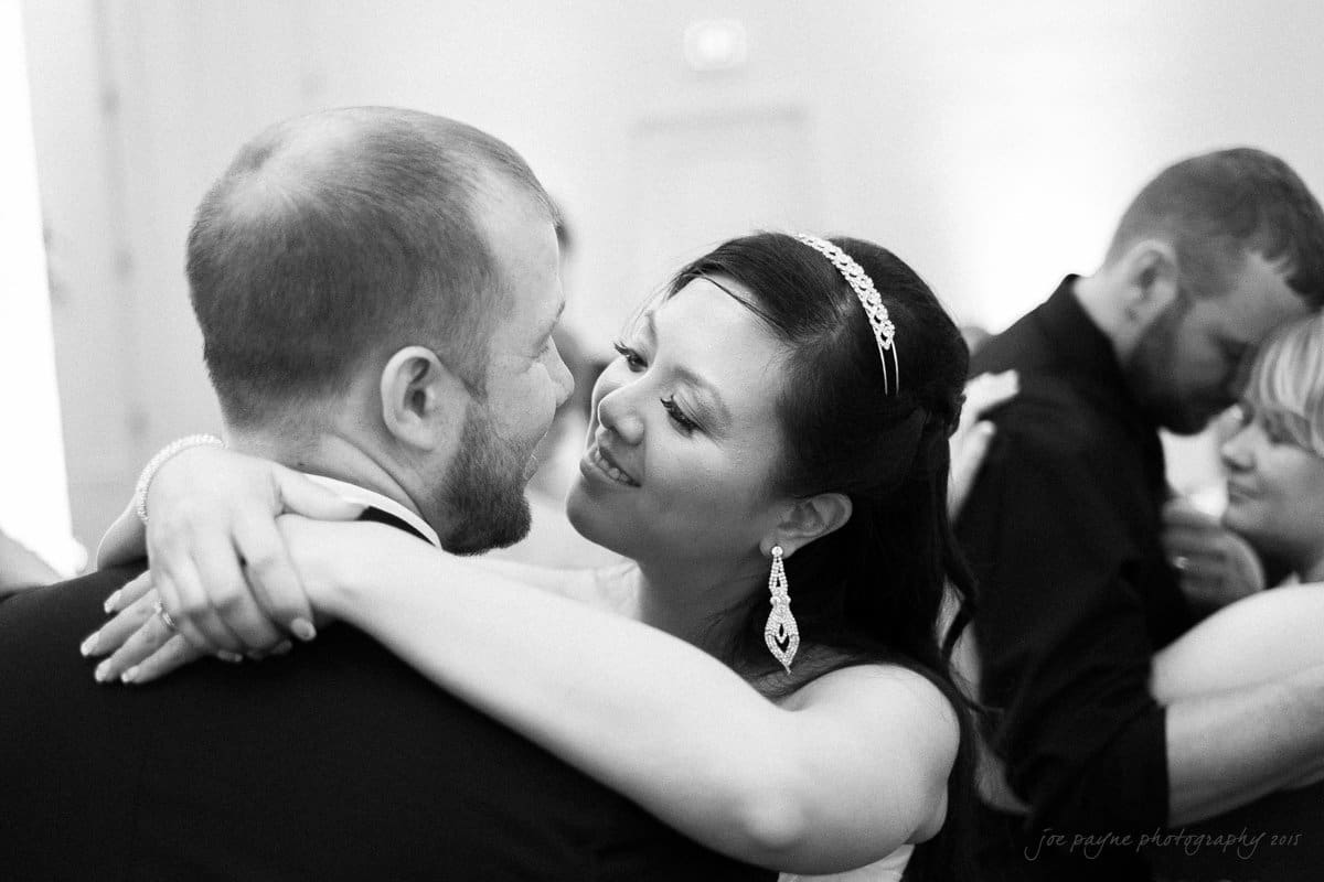 A bride and groom share a joyful embrace and smile at each other on the dance floor. They are surrounded by other guests in a softly lit room. The bride wears a tiara and earrings, while the groom is in a suit.