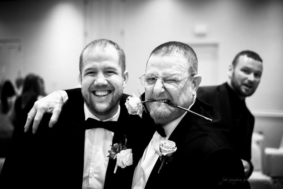 Three men in suits pose playfully at a formal event. Two of them stand side by side, with the older man holding a rose in his mouth and the younger grinning. Another man peeks over their shoulders, smiling.